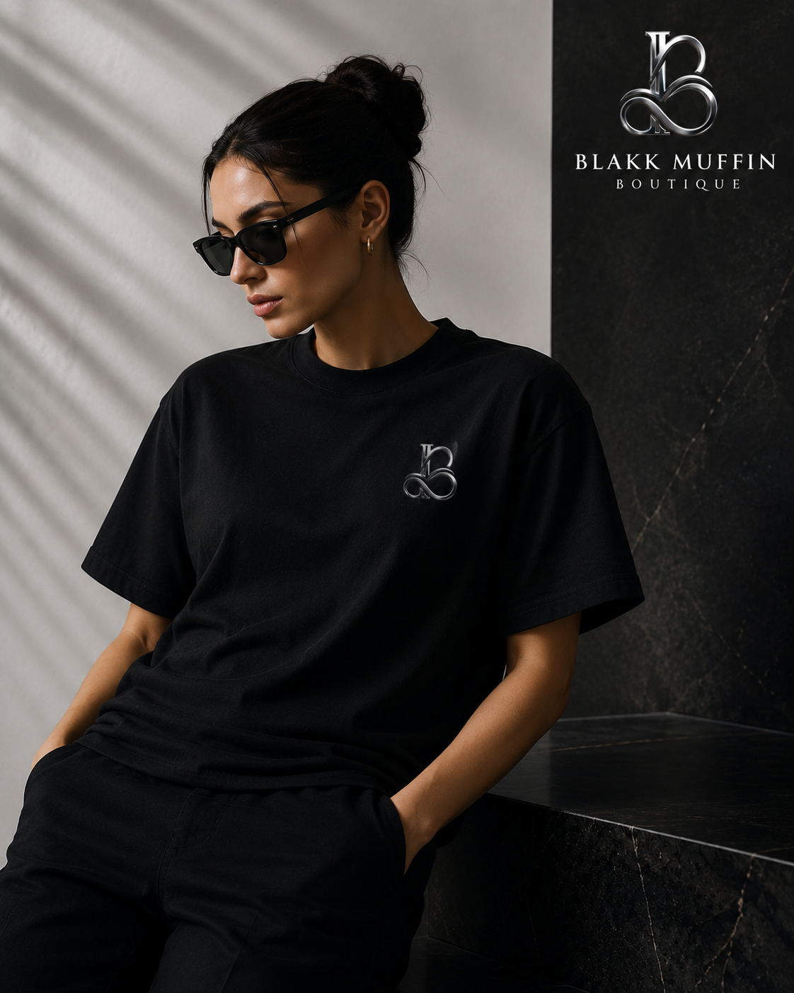 Woman wearing a black t-shirt with a logo, sitting against a gray wall with 'Blakk Muffin Boutique' branding.