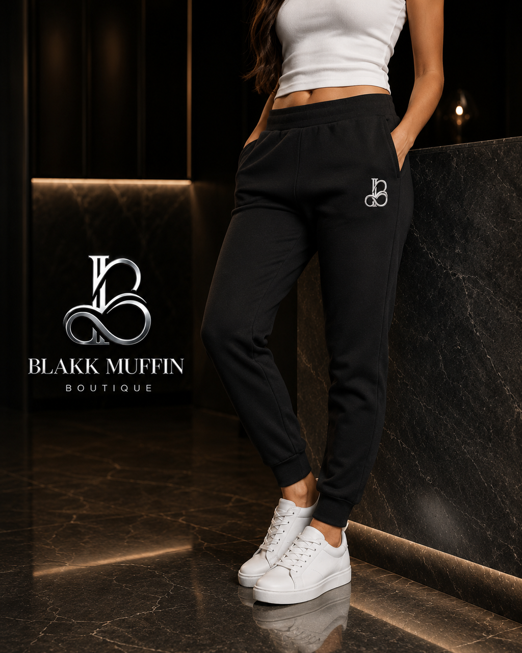 Person wearing black jogger pants with a logo and white sneakers on a dark background, featuring Blakk Muffin Boutique branding.